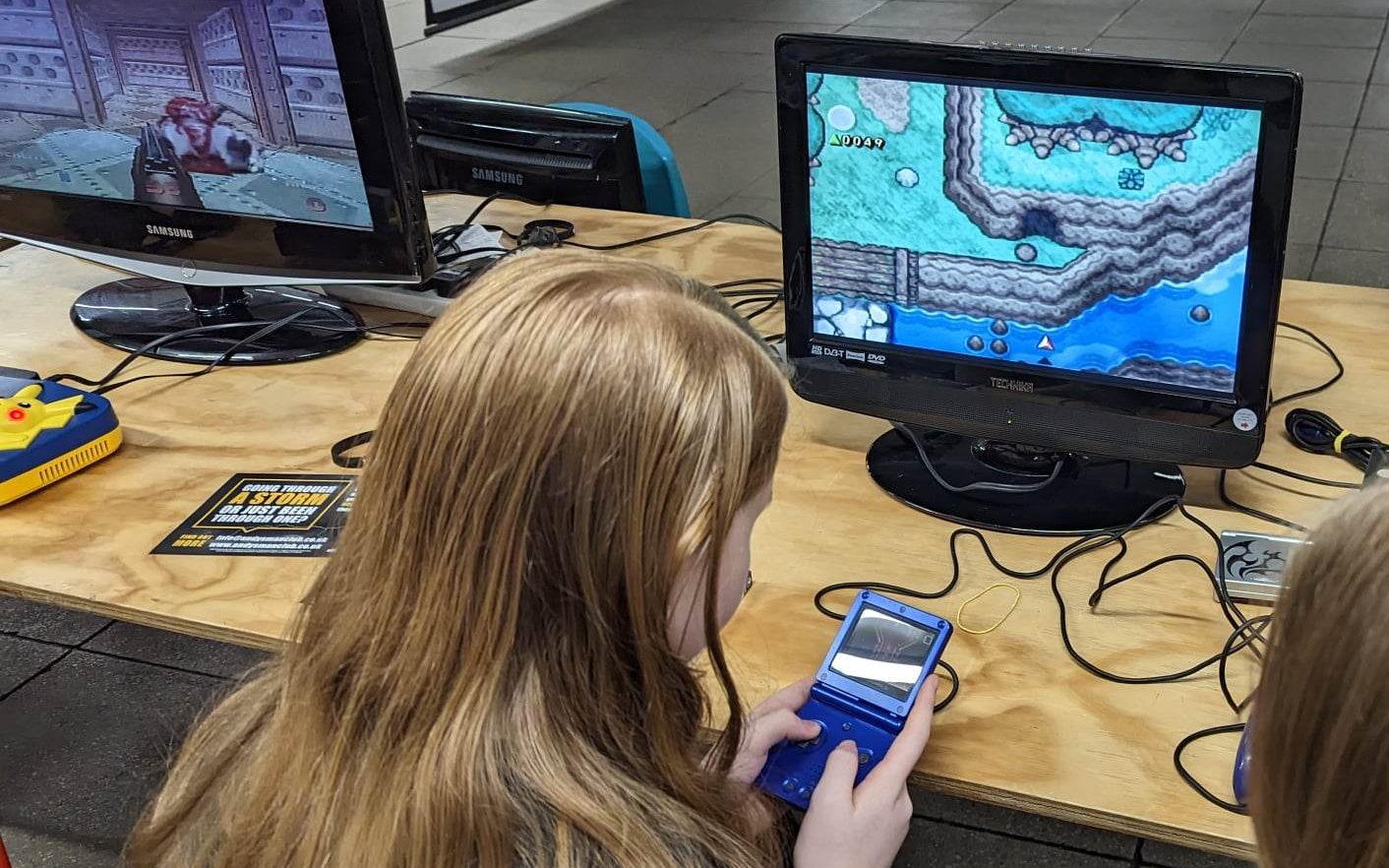 A girl with long hair playing a 'GameBoy Advance SP' connected to a TV screen.