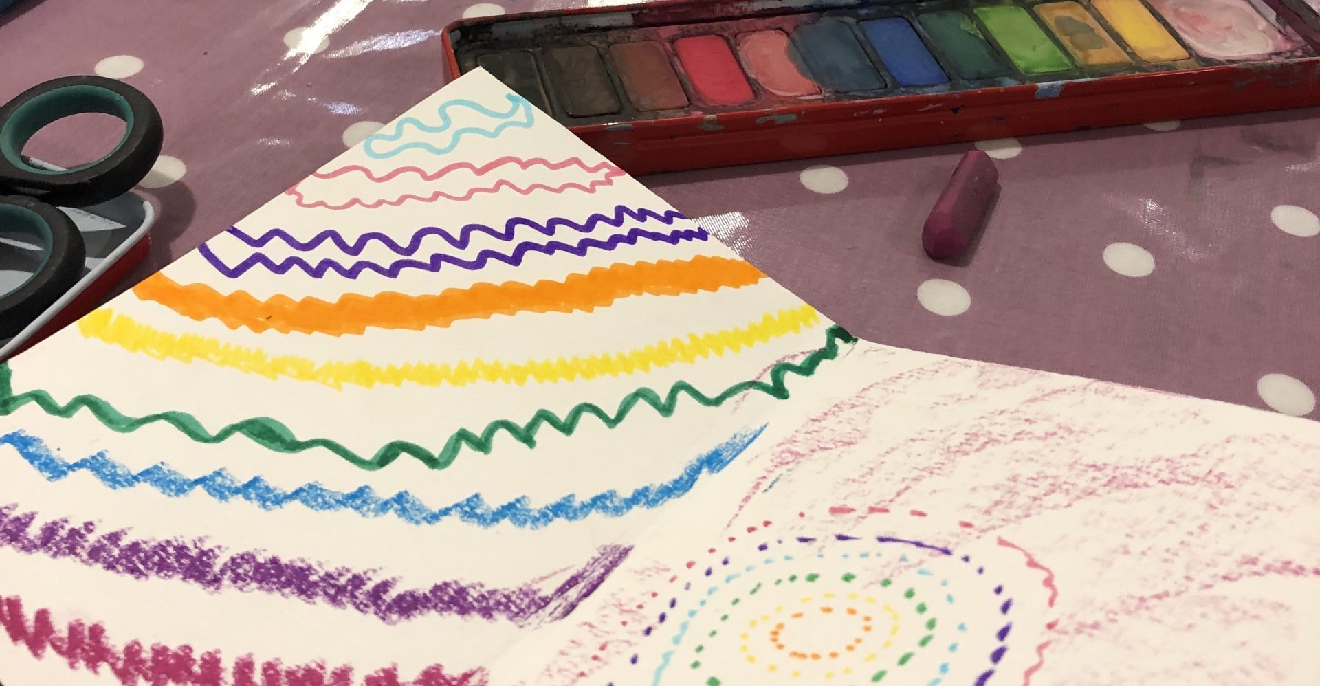 A zine (a paper booklet) decorated with colourful oil pastel patterns sits in the foreground. In the background art materials including a watercolour tablet and a pair of scissors can be seen.
