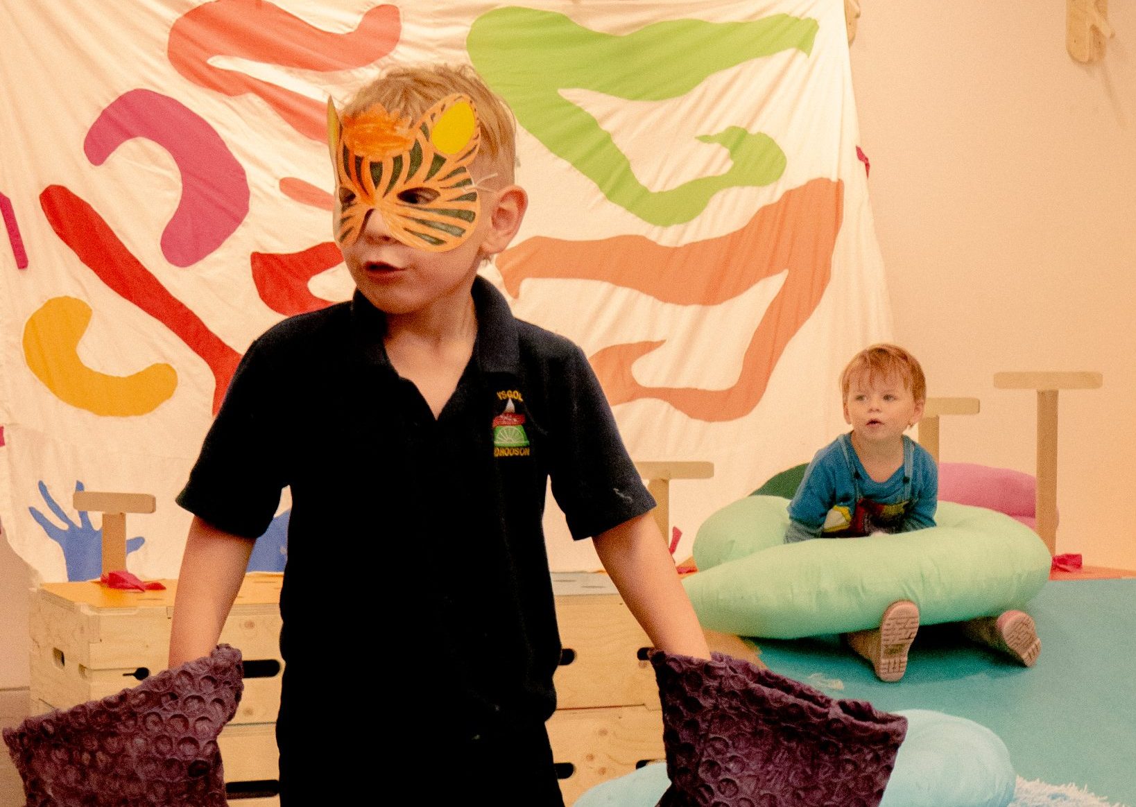 A child wearing a tiger mask, surrounded by colourful soft toys/artworks designed by Ella Jones. A toddler can be seen sitting in the background.