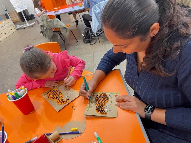 Family Art Club artist Manori demonstrates making a hedgehog to a primary school aged girl. They are sat together at an orange table in the Useful Art Space.