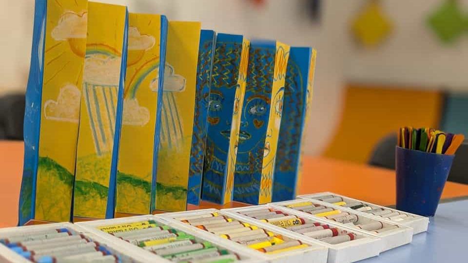 A blue and yellow artwork stands on a blue table, in front of it are boxes of oil pastels. In the background there is a dark blue cup of craft lolly sticks.