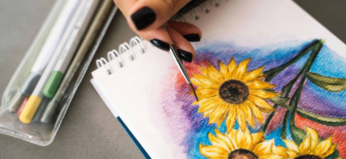An image of a hand with painted fingernails painting a watercolour of sunflowers. A clear pencil case of watercolour pencils can be seen to the left.