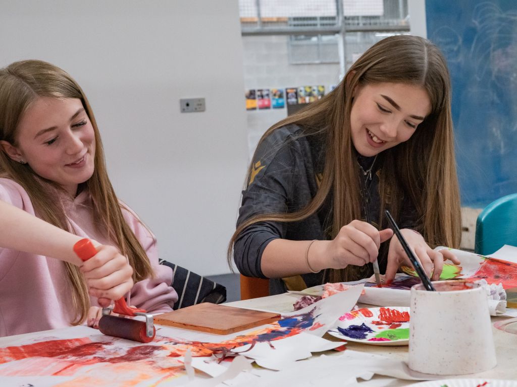 Two smiling teenagers work on their paintings. The girl on the left uses a roller. The girl on the right uses a small brush.
