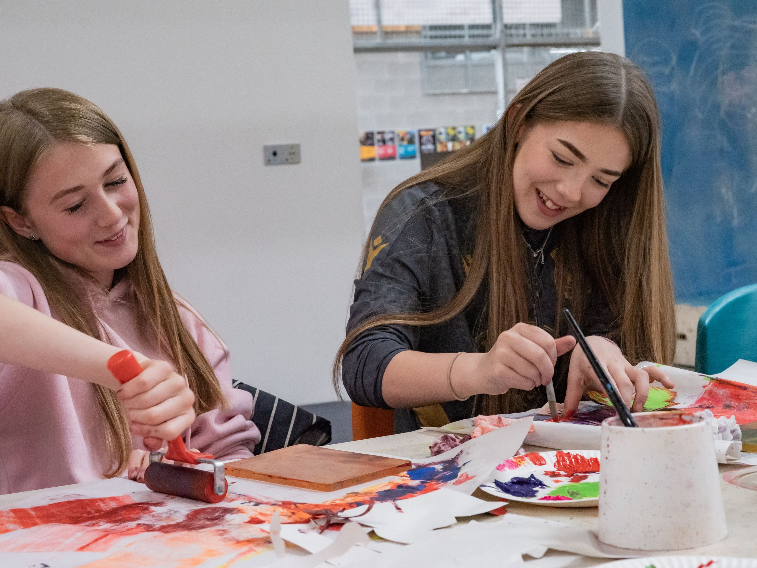 Two smiling teenagers work on their paintings. The girl on the left uses a roller. The girl on the right uses a small brush.