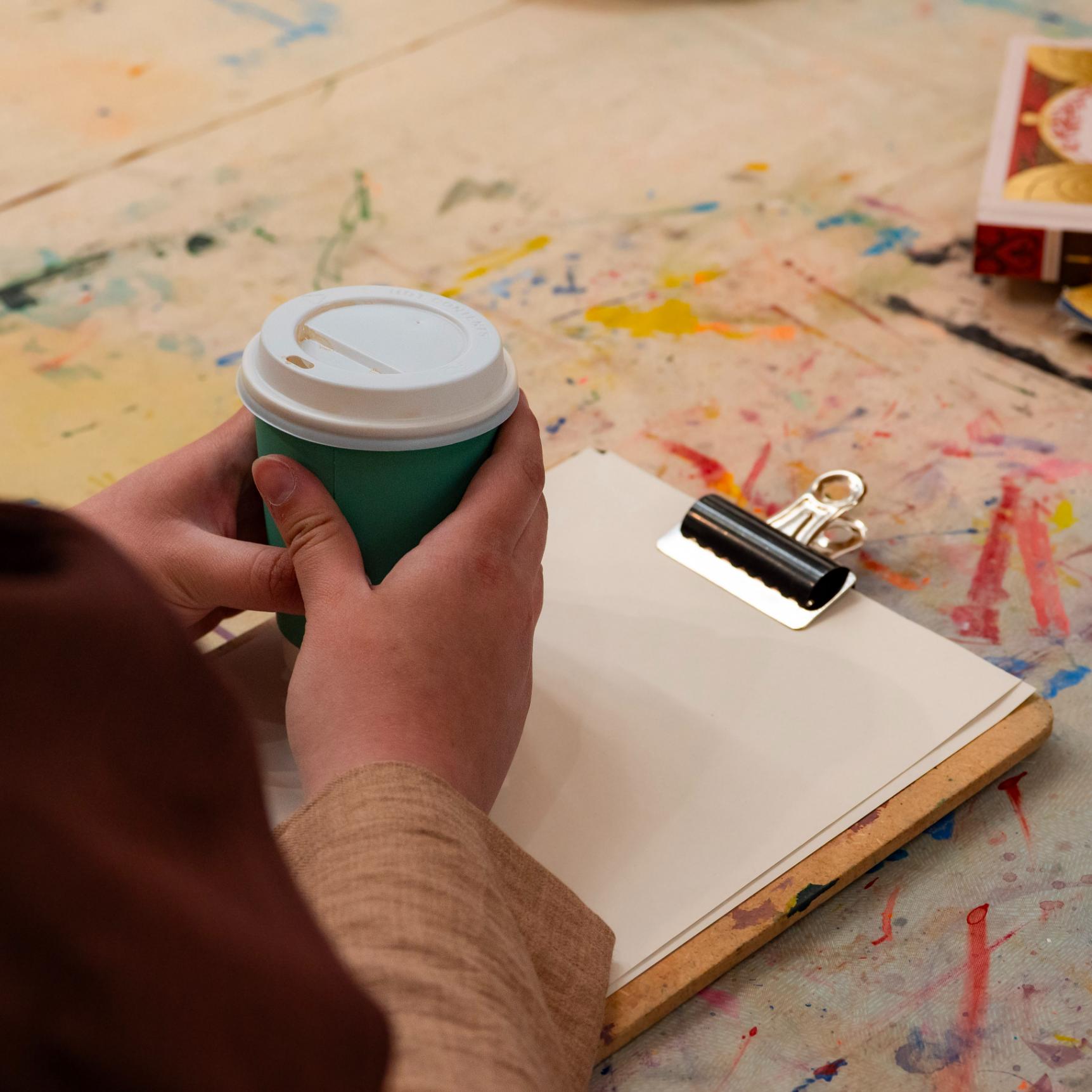 Hands holding a take-away cup of coffee. There is a clipboard with drawing paper and paint splattered table cloth on the table the person is sitting at.