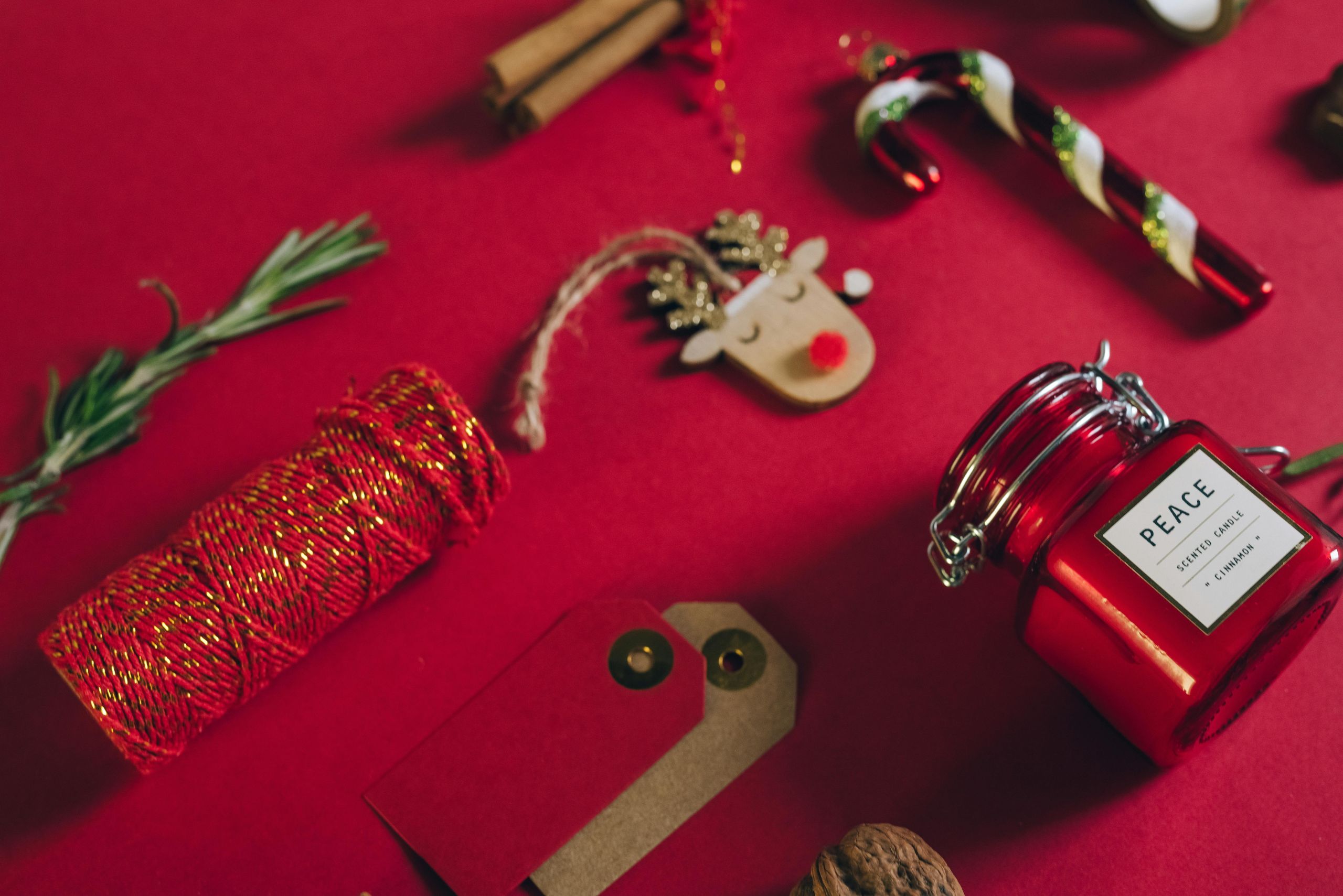 Christmas decorations and gift wrap supplies including Rudolph the Reindeer, a spool of red thread, paper gift tags, cinnamon sticks and a candle in a jar are laid out on a red table background.
