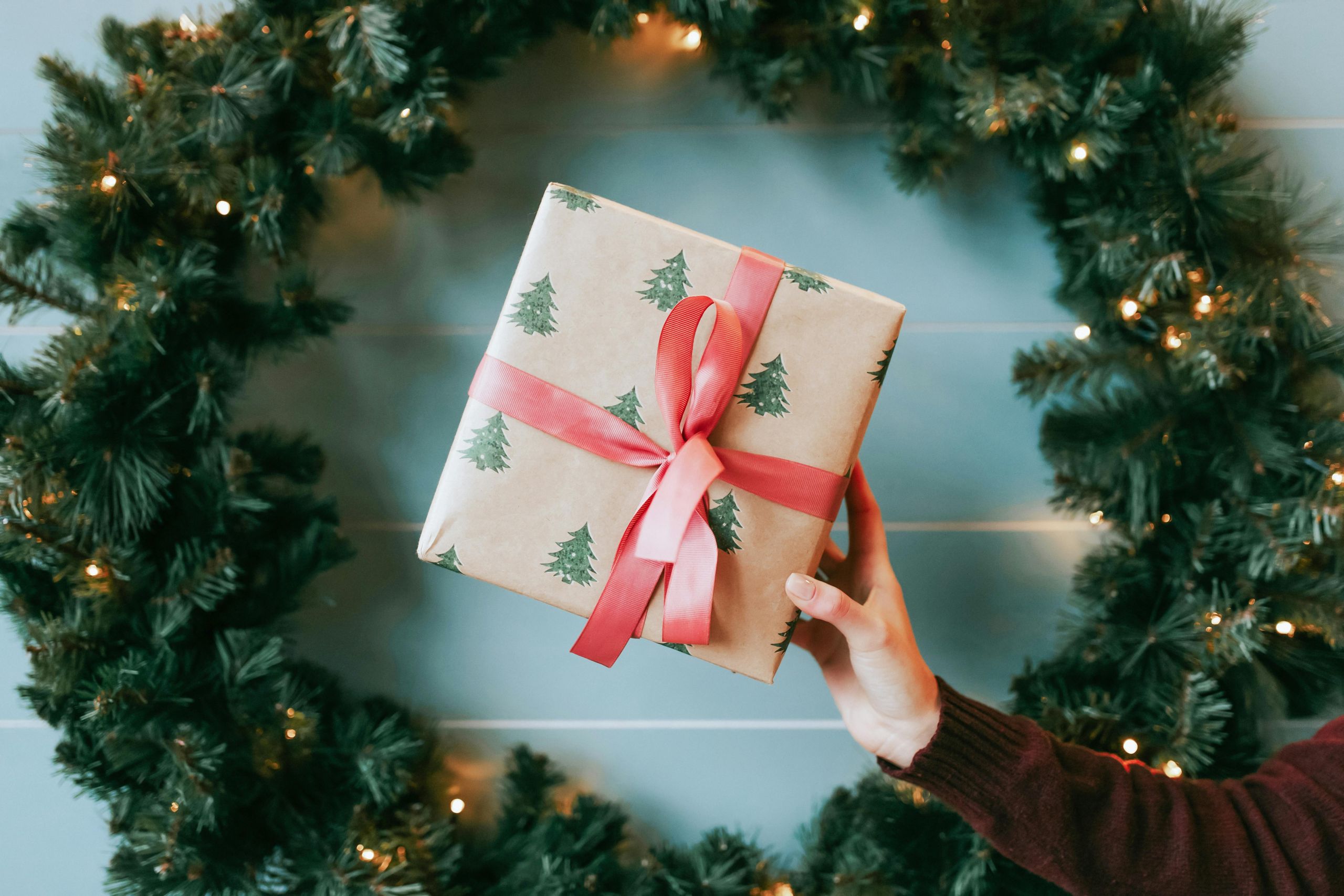 A hand holding a present wrapped in brown paper tied with a ribbon, surrounded by a twinkling Christmas wreath.