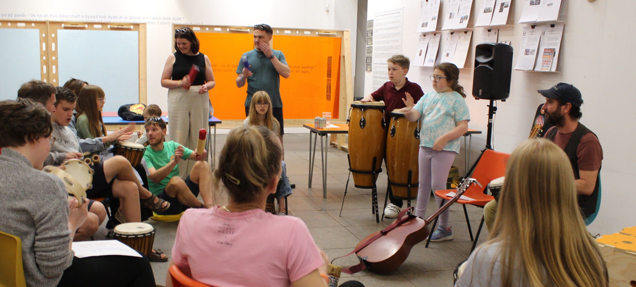 A family music workshop taking place. Children and adults can be seen playing drums in the Useful Art Space.