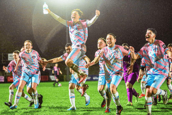 A football team dressed in trans flag colours celebrate on a brightly lit football pitch at night.