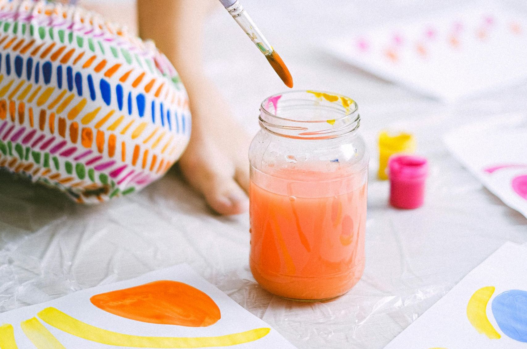A paintbrush being dipped into a jar of bright orange water by a young person. Their knee can be seen where they are kneeling on the floor to paint. They are wearing leggings with a blue, orange and yellow pattern. Around them on the floor are in progress paintings in yellow orange and blue drying out.