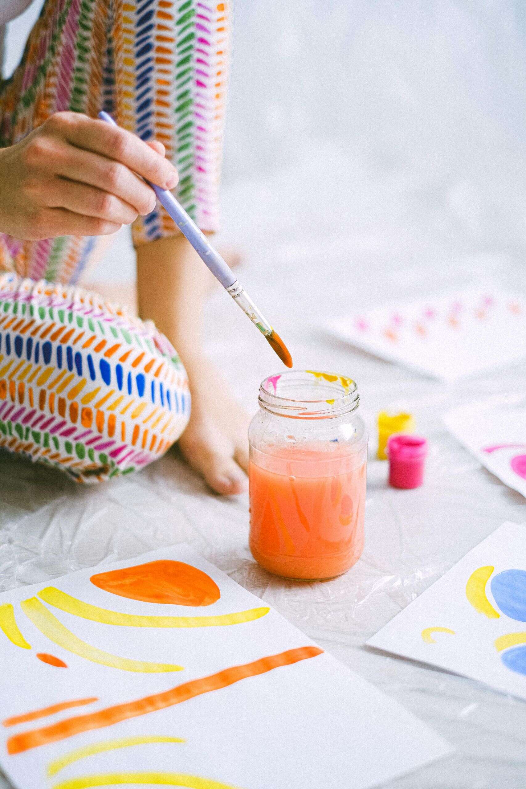 A paintbrush being dipped into a jar of bright orange water by a young person. Their knee can be seen where they are kneeling on the floor to paint. They are wearing leggings with a blue, orange and yellow pattern. Around them on the floor are in progress paintings in yellow orange and blue drying out.