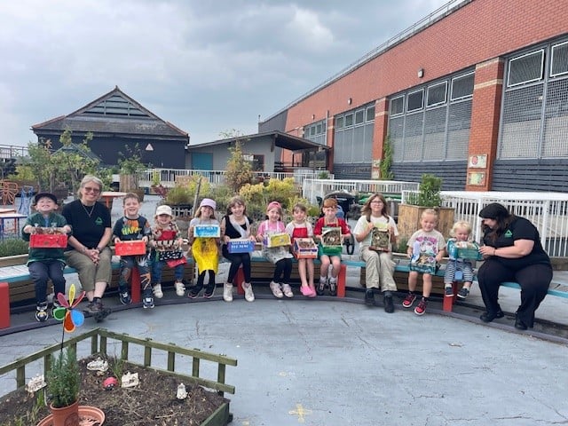A group of 12 children and adults are sat on the long, curved bench in our rooftop garden.