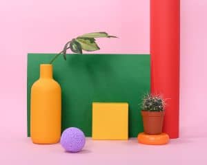 A still life composition featuring a yellow vase holding a single small branch, a purple ball (about the size of a tennis ball), and a short chunky cactus.