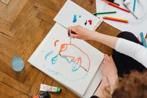 A hand working on a painting of a blue and orange face is seen from above.