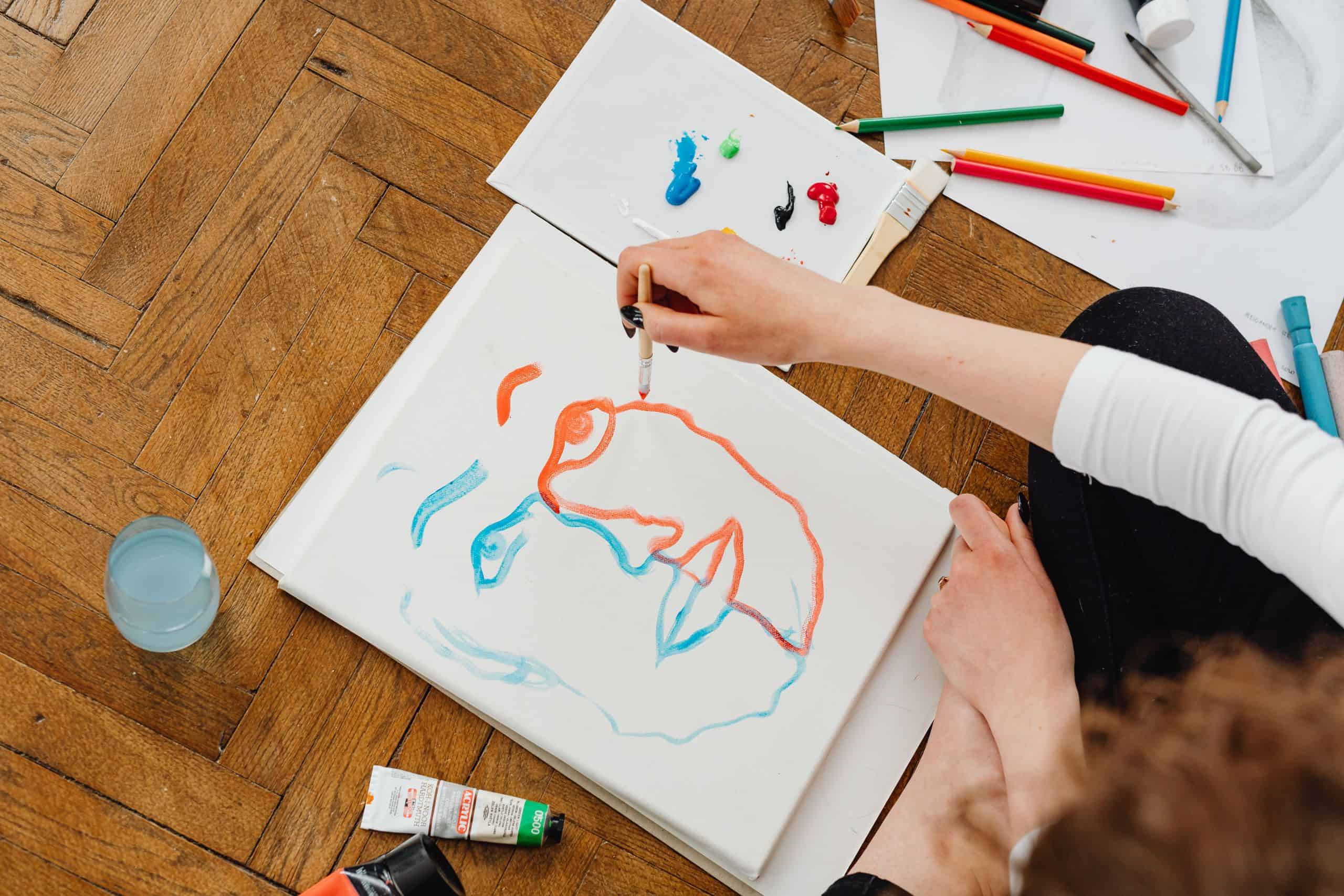 A hand working on a painting of a blue and orange face is seen from above.