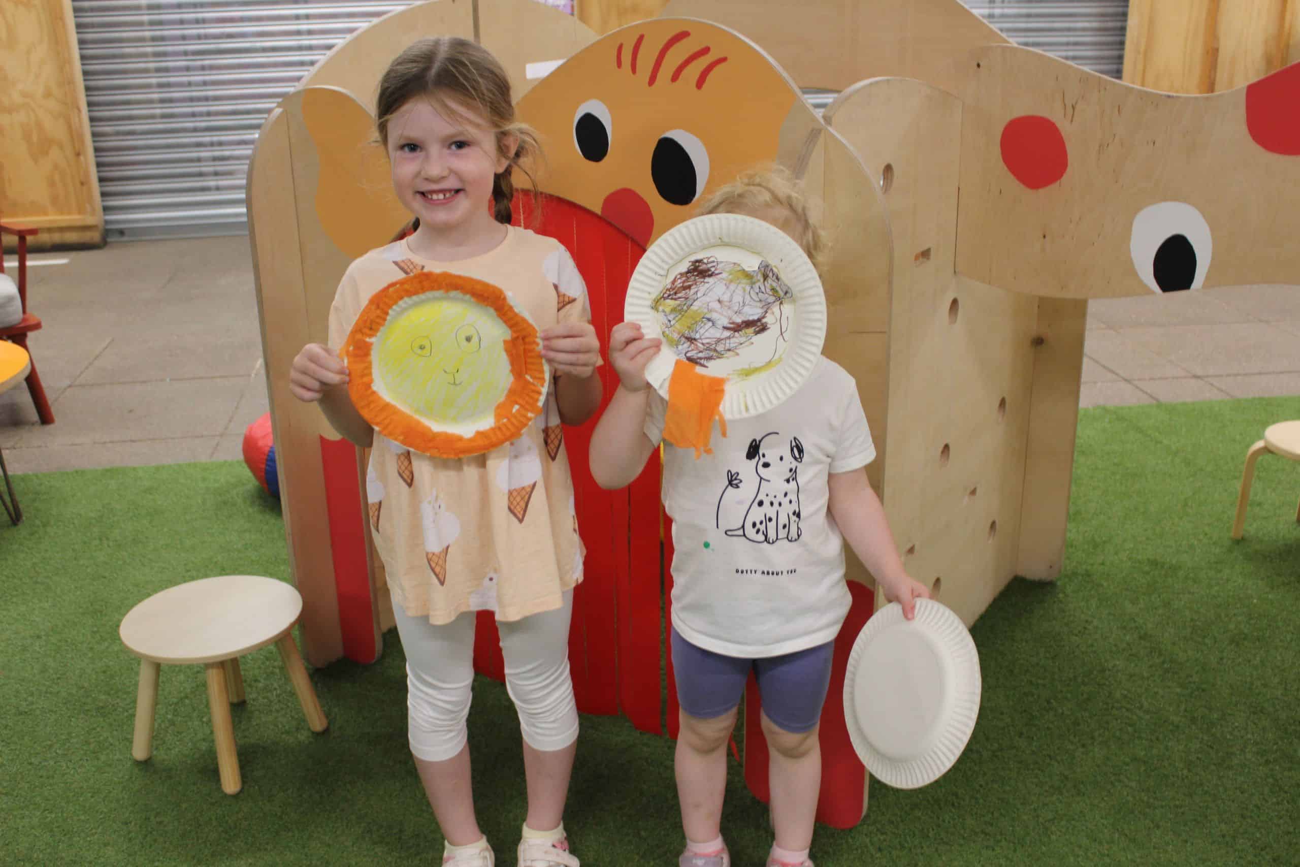 Two girls pose with the paper plate lion artwork they have made. Behind them is our indoor play equipment.