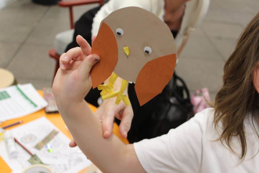 A child's hand holds up a collaged owl with brown paper wings, googly eyes and yellow paper straw legs.