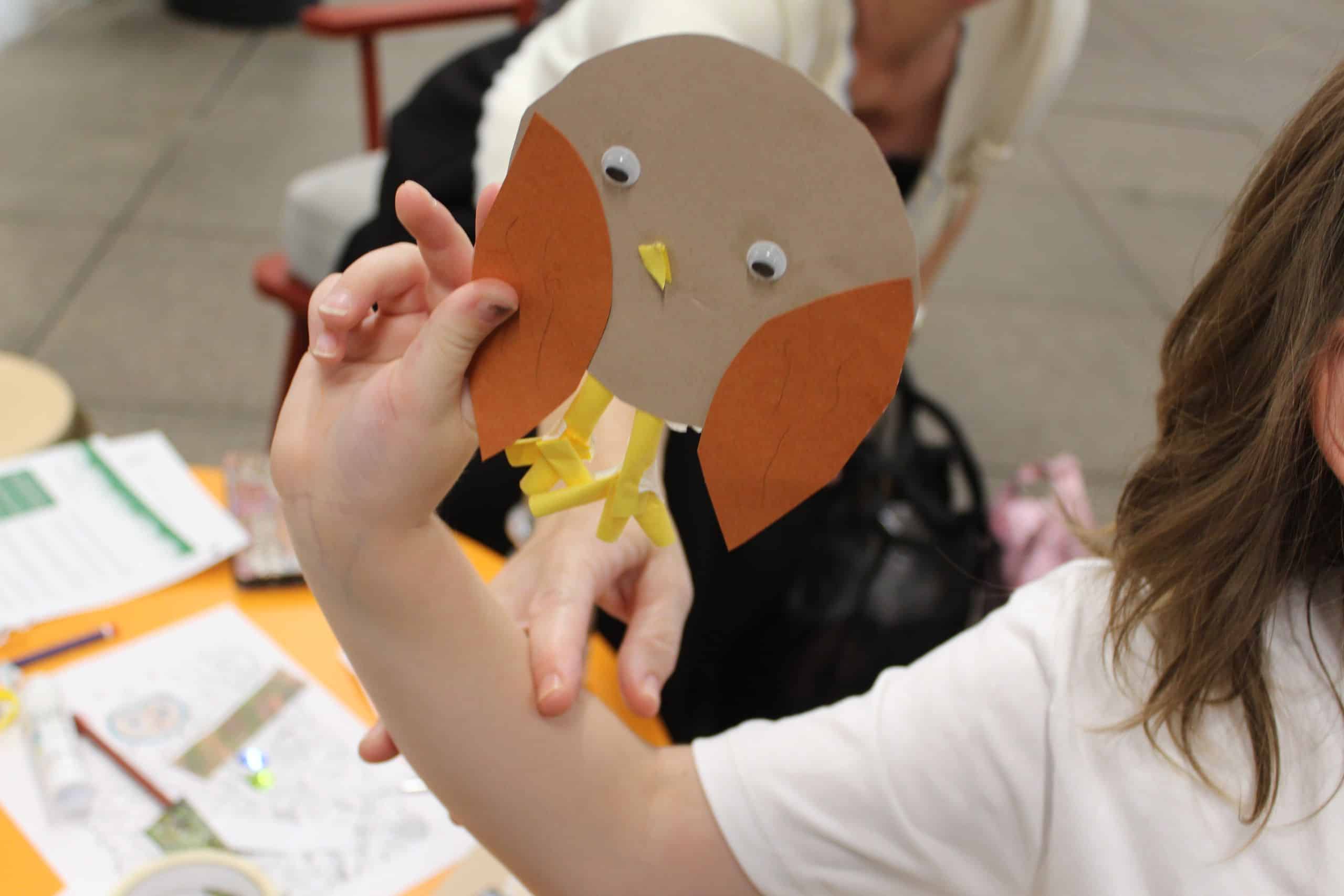 A child's hand holds up a collaged owl with brown paper wings, googly eyes and yellow paper straw legs.